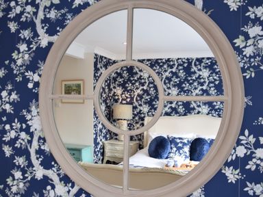 A bedroom with floral wallpaper a bed with blue and white cushions a bedside table with a lamp and a tray with cups at Napier House Southwold