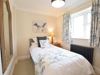 A single bed with white and blue floral bedding next to a window with matching curtains and a wooden nightstand with a lamp in a bedroom at Napier House Southwold