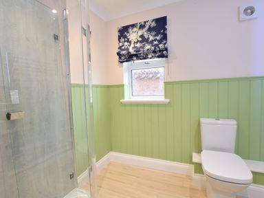 A bathroom with a glass shower enclosure a white toilet a window with a blue floral blind and green wainscoting at Napier House Southwold