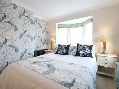 A bedroom with a double bed featuring peacock patterned bedding two lamps on bedside tables and a bay window with blinds at Napier House Southwold