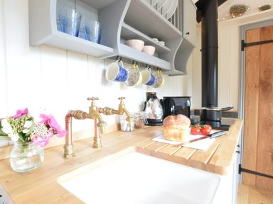 A kitchen countertop with a loaf of bread and tomatoes next to a sink with brass taps and shelves with mugs and bowls above at Shepherds Pightle in Hollesley