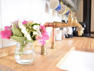 A kitchen countertop with vintage brass taps a glass jar with flowers and mugs hanging on a rack at Shepherds Pightle in Hollesley