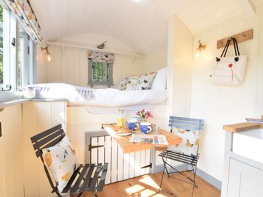A small room with a raised bed a wooden table set for breakfast two metal chairs with cushions and a window at Shepherds Pightle in Hollesley
