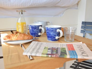 A wooden table set with two blue and white mugs, two glasses, a small jug of orange juice, croissants on plates, and a newspaper at Shepherds Pightle in Hollesley