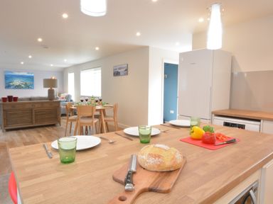 A kitchen island with a loaf of bread and cut bell peppers in a modern open plan kitchen and dining area with a wooden dining table set with plates and glasses and a living room with a sofa and a large painting at Bramertons Nest Long Melford