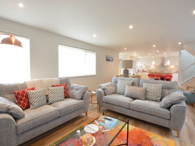A living room with two gray sofas and colorful pillows a glass coffee table and an open kitchen with red bar stools at Bramertons Nest Long Melford