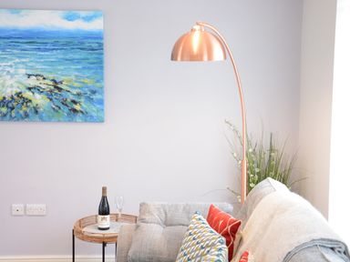 A corner of a living room with a gray sofa with cushions and a blanket a copper floor lamp a side table with a bottle of wine and a glass a blue ocean painting on the wall and a plant in the corner at Bramertons Nest Long Melford