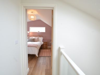 A bedroom with a wooden bed frame pillows a bedside table and a rug viewed from a hallway at Bramertons Nest Long Melford
