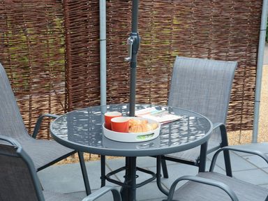 An outdoor patio with a round glass table and four grey chairs with a tray holding two orange cups and croissants at Bramertons Nest Long Melford