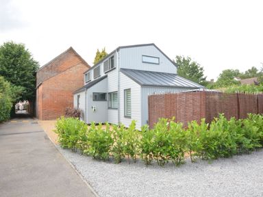 A modern house with gray siding next to a brick building and a gravel driveway with green bushes at Bramertons Nest Long Melford