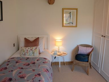 A bedroom with a single bed a side table with a lamp a cup and a magazine and a wicker chair with cushions at May Barn Ixworth in Bury St Edmunds