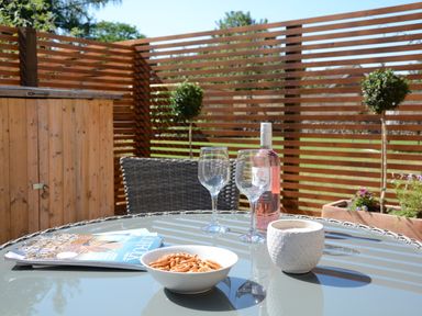 A glass outdoor table with a bowl of pretzels a white cup two wine glasses a bottle of rose wine and magazines in a patio area with wooden fencing and plants at May Barn in Ixworth