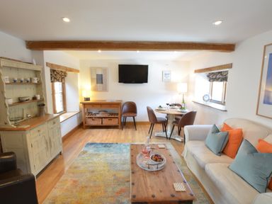 A living room with a beige sofa with blue and orange pillows a wooden coffee table a dining table with four chairs two windows a wooden cabinet and a flat screen tv on the wall at May Barn Ixworth in Bury St Edmunds