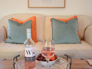 A beige sofa with blue and orange cushions and a wooden table with a bottle of pink wine a glass of wine and a bowl of fruit at May Barn Ixworth in Bury St Edmunds