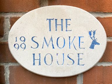 An oval stone plaque with blue carved text reading The Smoke House and the year 1899 on a brick wall at The Smokehouse Cottage in Southwold