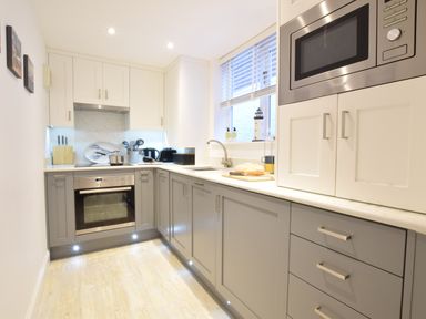 A kitchen with gray lower cabinets white upper cabinets an oven microwave a sink and a window at The Smokehouse Cottage in Southwold