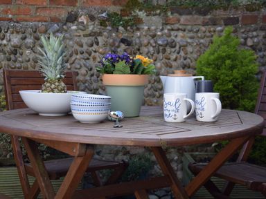 A wooden outdoor table with a bowl containing a pineapple two patterned bowls a green flower pot with flowers a white teapot two white mugs and a spoon at The Smokehouse Cottage in Southwold