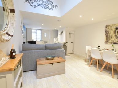 A living area with a grey sofa a wooden storage chest a dining table with white chairs and a mirror on the wall at The Smokehouse Cottage in Southwold