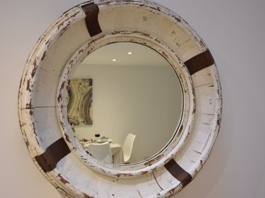 A round wall mirror with a distressed white wooden frame reflecting a dining area with white chairs and a table at The Smokehouse Cottage in Southwold