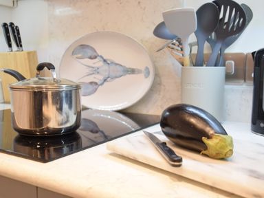 A kitchen countertop with a stainless steel pot on a stove an eggplant and knife on a cutting board a knife block a plate with a lobster design and utensils in a holder at The Smokehouse Cottage in Southwold