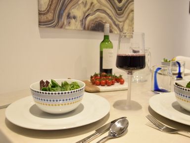 A dining table set with bowls of salad on plates a glass of red wine a bottle of wine and cherry tomatoes on a cutting board at The Smokehouse Cottage in Southwold