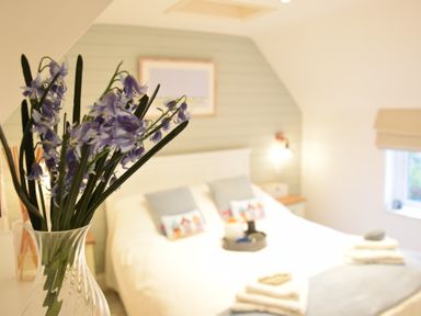 A bedroom with a bed with pillows and folded towels a window and a vase with purple flowers in the foreground at The Smokehouse Cottage in Southwold