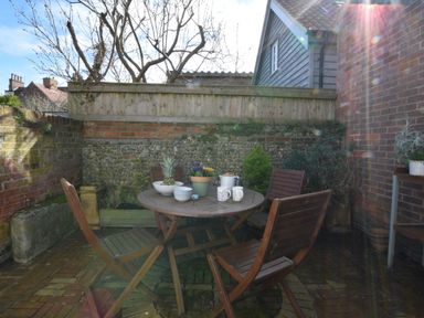 An outdoor patio with a round wooden table and four wooden chairs against a brick and stone wall at The Smokehouse Cottage in Southwold