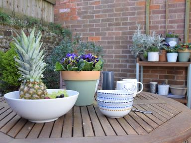 A wooden patio table with a bowl of fruit a flower pot and cups in a garden at The Smokehouse Cottage Southwold