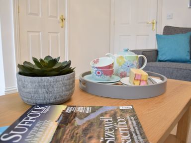 A coffee table with a succulent plant, a magazine, and a tray holding a teapot, two teacups, and a piece of cake in a living room with a gray sofa and blue cushion at Bluebelle Cottage Peasenhall