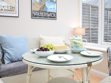 A glass top table with plates cutlery a bowl of grapes cheese a teapot and cups by a sofa with cushions in a room with windows and framed pictures at Bluebelle Cottage Peasenhall