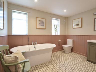 A bathroom with a freestanding bathtub a mint green chair with pillows a toilet framed pictures on walls and two windows at Satis Cottage Westleton
