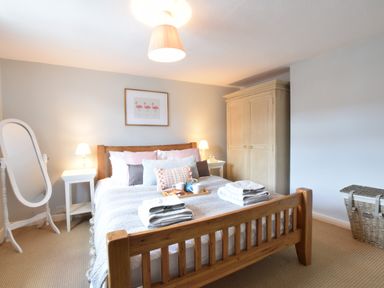 A bedroom with a wooden bed two bedside tables with lamps a wardrobe a standing mirror and a wicker basket at Satis Cottage Westleton