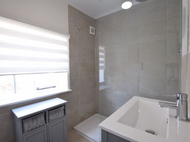 A bathroom with a sink and faucet a shower area a window with a blind and a cabinet with baskets at Satis Cottage Westleton