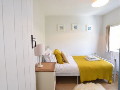 A bedroom with a bed covered with a yellow blanket and pillows a bedside table with a lamp a window with curtains and three framed pictures on the wall at Satis Cottage Westleton