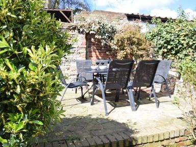 An outdoor patio with a glass table and black chairs surrounded by greenery and a brick wall at Satis Cottage Westleton