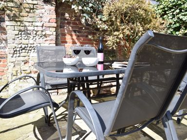 A glass table with two wine glasses bowls of fruit and a bottle on a patio with metal chairs and a brick wall at Satis Cottage Westleton