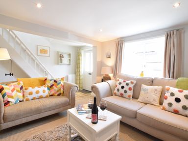 A living room with two sofas, colorful cushions, a white coffee table with wine and glasses, and a staircase in the background at Satis Cottage, Westleton