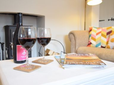 A table with two glasses of red wine a bottle of wine magazines a beige sofa with colorful cushions and a fireplace in the background at Satis Cottage Westleton