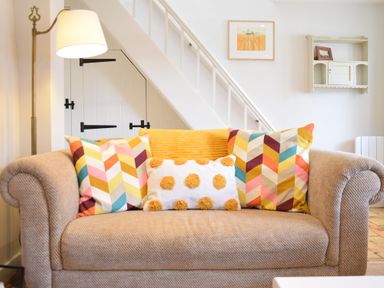 A beige sofa with colorful patterned cushions and a lamp beside it under stairs at Satis Cottage Westleton