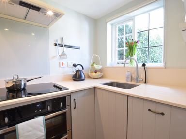 A kitchen corner with a stove, kettle, fruit basket, sink, and window with flowers at Satis Cottage Westleton