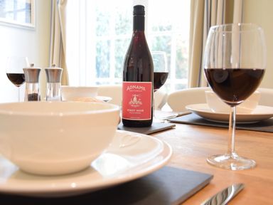 A dining table set with white bowls and plates red wine glasses a bottle of Adnams Pinot Noir and salt and pepper shakers at Satis Cottage Westleton