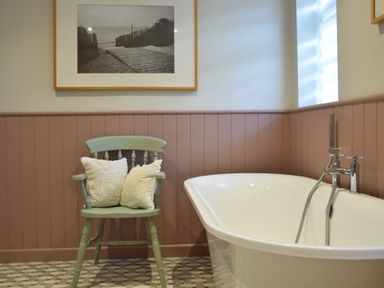 A bathroom with a white freestanding bathtub a green wooden chair with two cushions and a framed black and white photograph on the wall at Satis Cottage Westleton