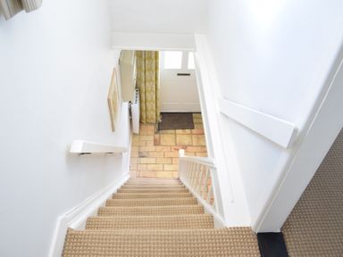 A staircase with beige carpet leading down to a tiled entryway with a white door and yellow patterned curtain at Satis Cottage Westleton