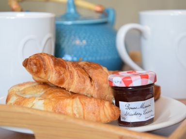 A breakfast tray with croissants a jar of strawberry jam two white mugs and a blue teapot at Satis Cottage Westleton
