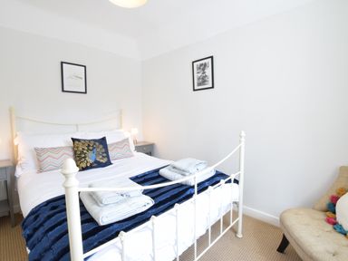 A bedroom with a white metal bed frame two bedside tables two framed pictures on the walls and a beige upholstered chair at Satis Cottage Westleton