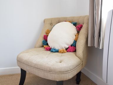 A beige upholstered chair with a round white cushion with colorful tassels near a window with curtains at Satis Cottage Westleton