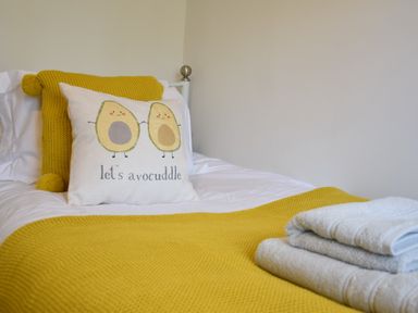 A bed with a yellow knitted throw and pillow and a decorative pillow with avocado design and text at Satis Cottage Westleton