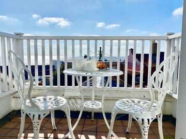 A small balcony with a white metal table and two chairs with teapot cups and pastries overlooking rooftops and the sea at Albion House Southwold