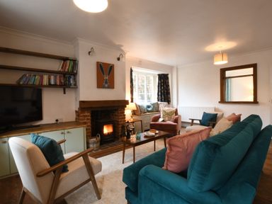 A living room with a teal sofa pink and teal cushions armchairs a wooden coffee table a fireplace with a fire a window seat with cushions and shelves with books at Rosedene Rickinghall