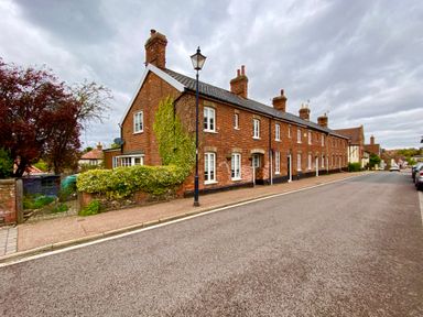 A row of brick houses with chimneys and white windows along a street with a lamppost and parked cars at Rosedene Rickinghall